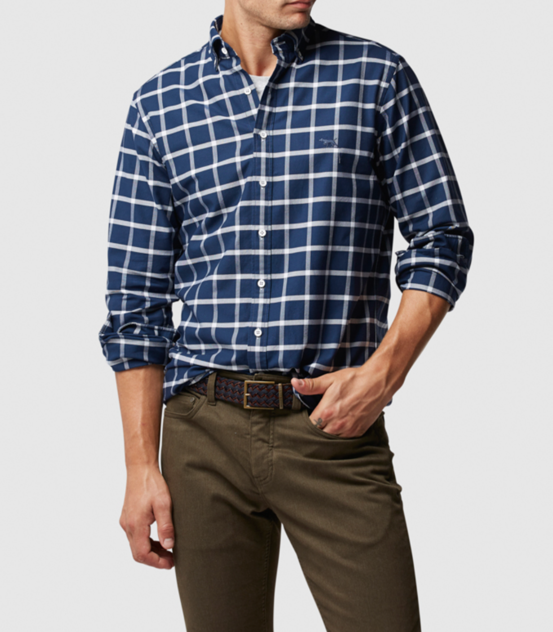 Wearing a Rodd & Gunn Gunn Check Oxford Sports Fit Shirt in blue and white checkered 100% cotton with rolled sleeves, and olive green pants, one hand rests in their pocket. A detailed belt stands out against a plain gray background, with the face not shown.