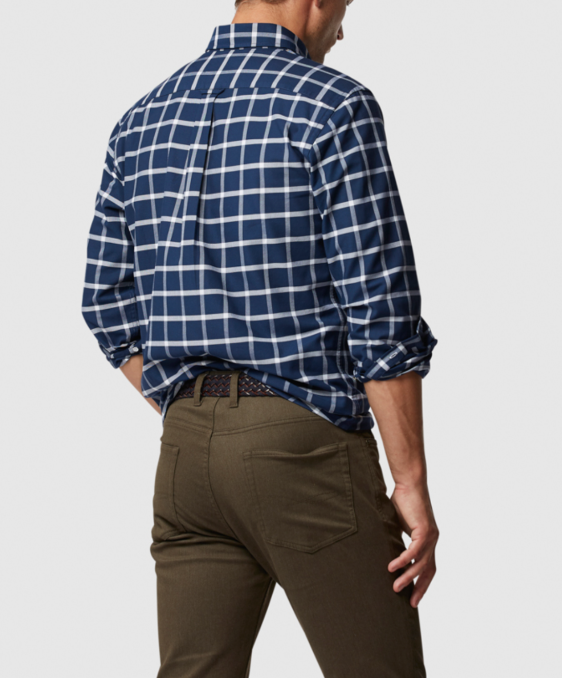 A person is seen from behind wearing a Rodd & Gunn Gunn Check Oxford Sports Fit Shirt in blue and white checks, crafted from 100% cotton, paired with olive green pants and a black belt. Their right hand rests on their hip, and their head is slightly turned to the right against a plain white background.