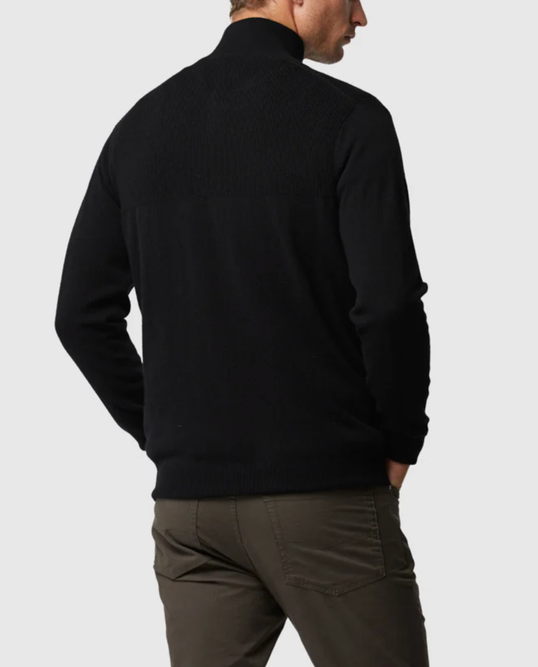 A person wearing the Rodd & Gunn Little Mount Allan Knit, a black Merino Cashmere blend zip-up sweater, along with khaki pants stands facing away with hands in pockets. The plain white background emphasizes the clothing.