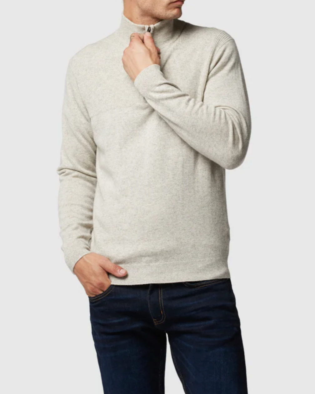 A person stands against a plain background wearing the Rodd & Gunn Little Mount Allan Knit, a light gray, long-sleeved Australian Optim Wool sweater with a short zipper at the neck. The individual has one hand pulling up the zipper and the other hand resting in a pocket of dark blue jeans.