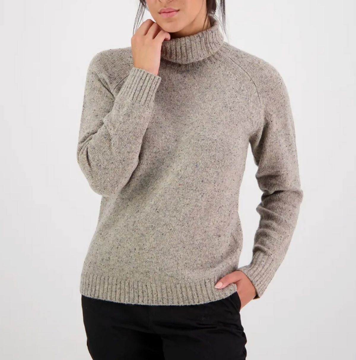 A person is wearing a Swanndri Lyttleton Roll Neck Jumper in light gray, crafted from lambswool, featuring long sleeves and ribbed cuffs. They are standing with one hand in the pocket of their black pants against a plain white background. The person has long, dark hair tied back.