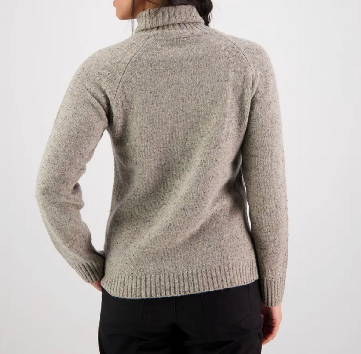 A person with long dark hair is seen from the back, wearing the stylish Swanndri Lyttleton Roll Neck Jumper in light gray lambswool with a textured, knitted appearance, paired with black pants. The background is a plain, light gray color.