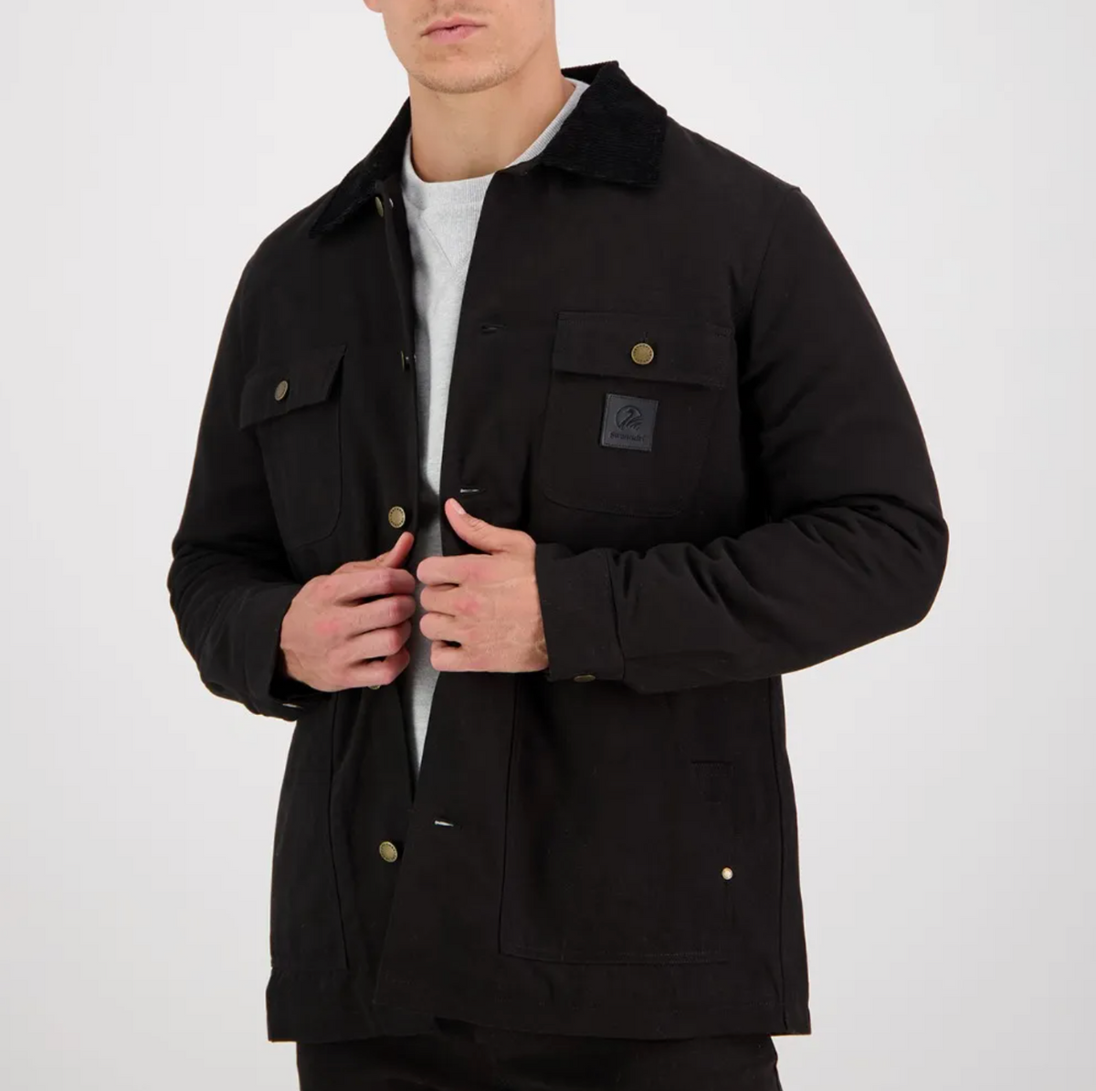 A person in a Swanndri Barrytown Canvas Work Jacket, featuring buttoned pockets and a black collar, is standing against a plain white background. They are wearing a light gray shirt underneath and are adjusting the jacket with their hands. The jacket, which features quilted lining, has a patch on the left chest pocket.