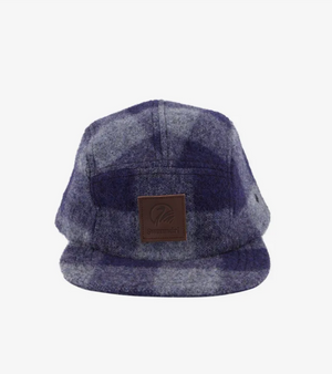 A front-facing view of the Swanndri Wentworth Valley Cap showcases its blue and gray plaid design. This five-panel cap, made of 100% wool, features a short, flat brim and a brown leather logo patch on the front panel. The background is plain white.