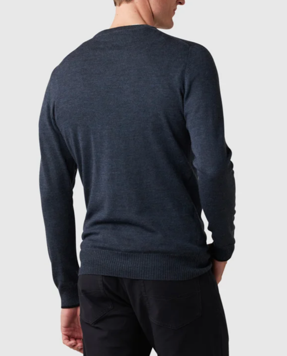 In a plain light gray setting, a person faces away wearing the Rodd & Gunn Abberfield Knit sweater in dark blue merino wool, paired with black pants. The sweater has long sleeves and a ribbed hem.