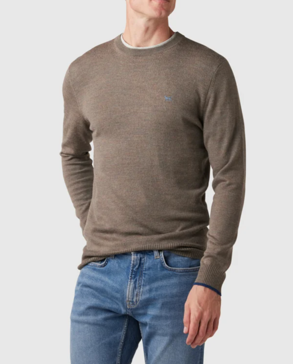 Dressed in a light brown Rodd & Gunn Abberfield Knit sweater with an embroidered chest logo, a person pairs it with blue jeans and stands against a white backdrop, hands tucked casually into their jean pockets.