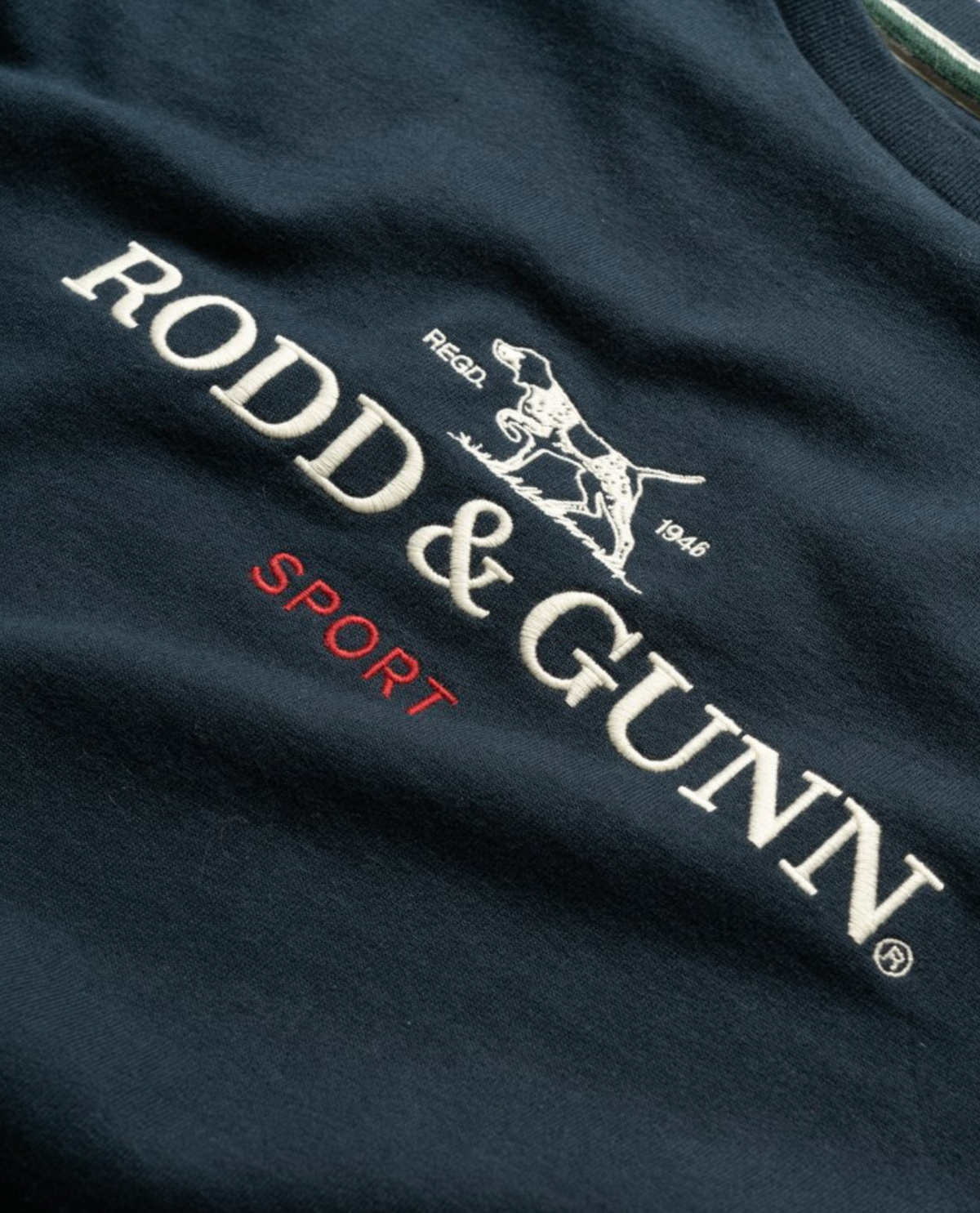 Close-up of the Rodd & Gunn Grenada North Sweat, a navy blue 100% cotton garment featuring the brand "Rodd & Gunn", a logo of a pointer dog, and the words "Regd. 1946" all in white stitching. The word "Sport" is embroidered underneath in red stitching, showcasing the stylish design of this comfortable sweat from Rodd & Gunn.