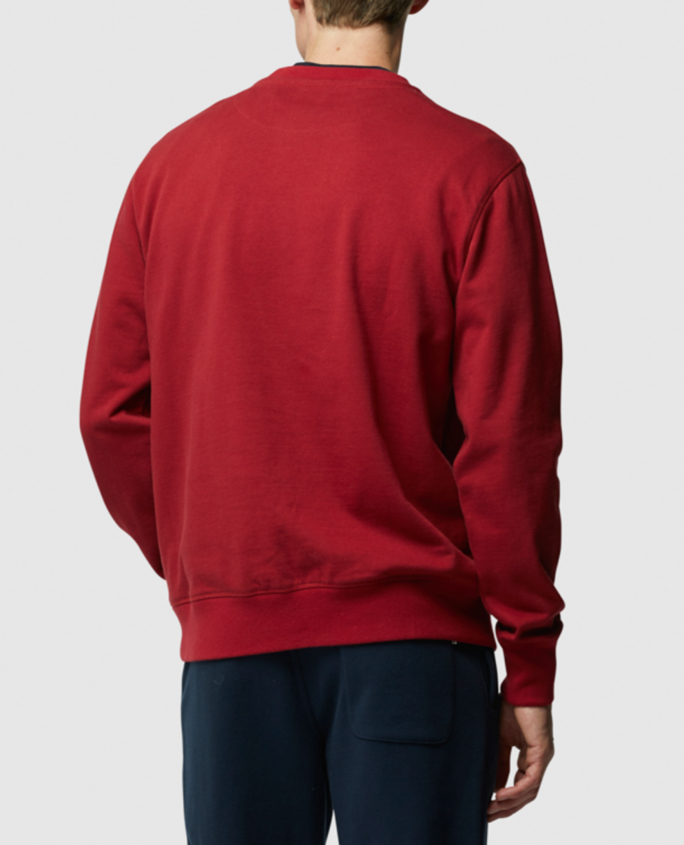 The image displays a person from behind wearing the Rodd & Gunn Grenada North Sweat, a plain red crew neck sweatshirt with subtle brand embroidery, paired with dark pants against a neutral background.