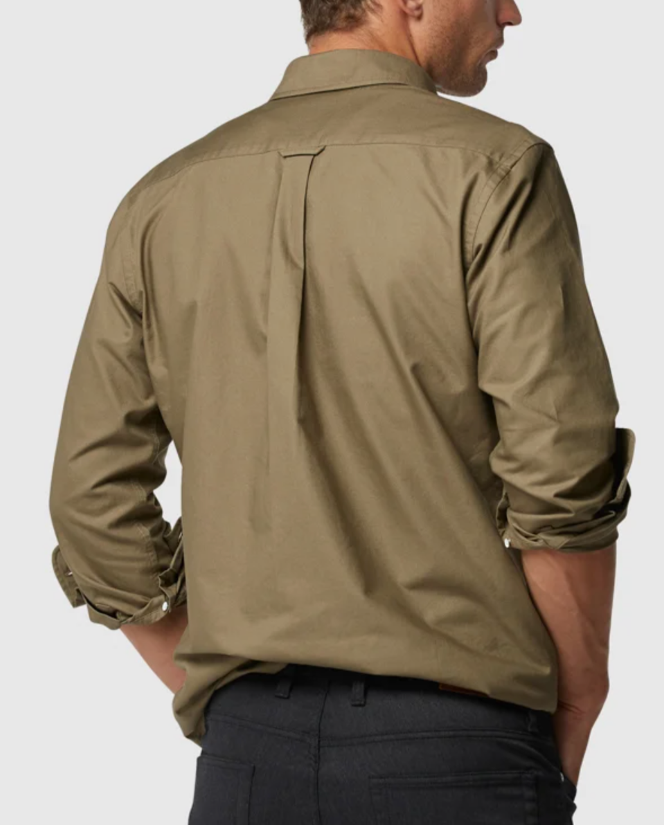 A man in a Rodd & Gunn Oxford Pd Sports Fit Shirt, light brown and 100% cotton, stands with sleeves rolled up and hands on hips, sporting dark pants. The pleat back design complements his posture as he faces a plain background.
