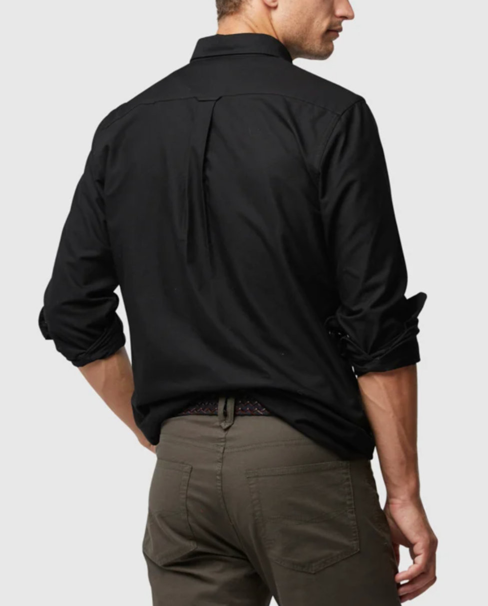 A person wearing a stylish Rodd & Gunn Oxford Pd Sports Fit Shirt in black, with rolled-up long sleeves, and comfortable brown pants stands with their back to the camera against a plain background.