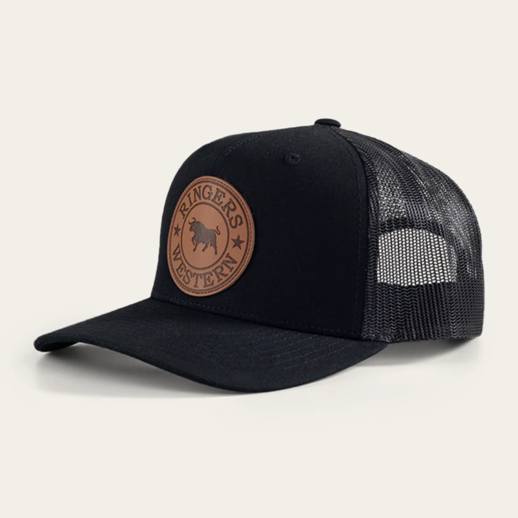The Ringers Western Whitby Trucker Cap features a black design with a brown leather patch showcasing the "Ringer's Western" text and bull logo. Its mesh back provides ventilation, while the curved brim and adjustable snap closure ensure comfort and style.