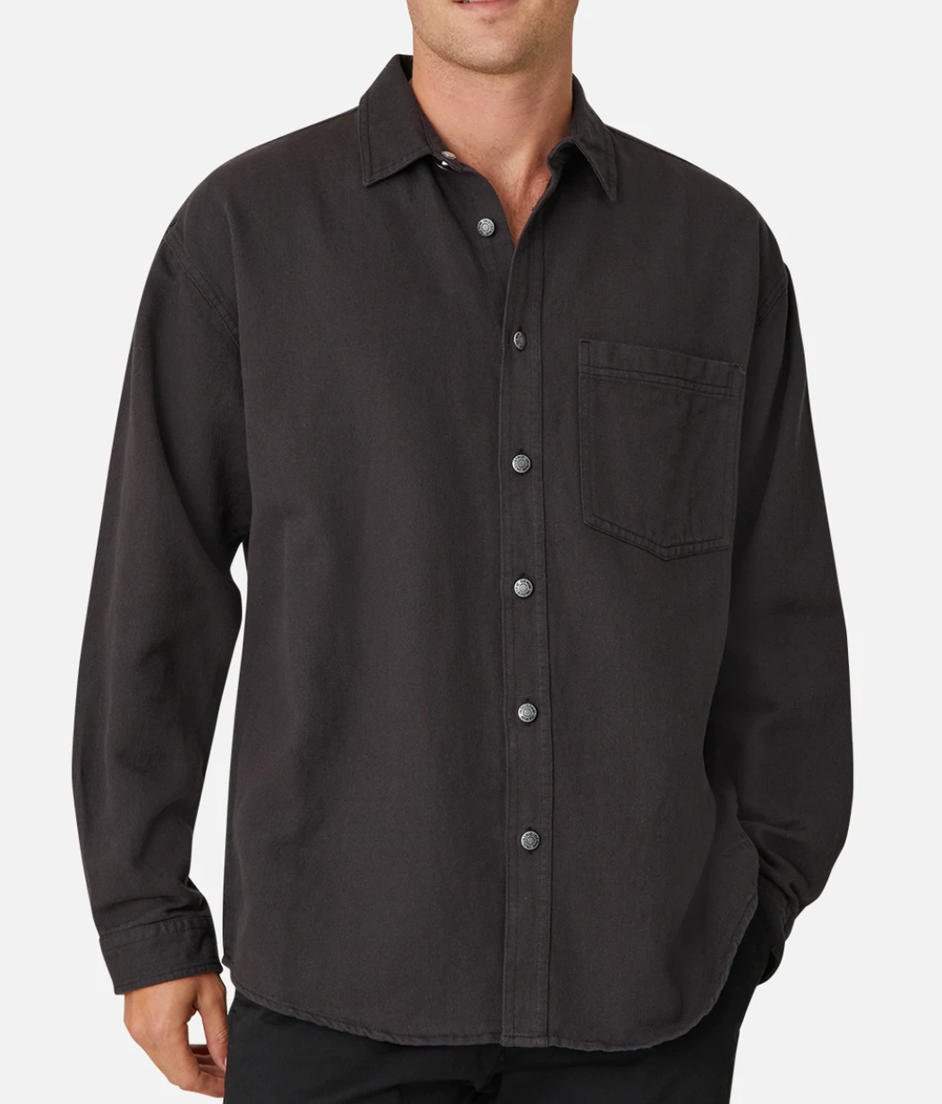 A person is wearing the Industrie The Portland Shirt, a long-sleeve, dark-colored button-up with a collar and a single chest pocket on the left side. The stylish shirt features an Industrie logo on the pocket and has a casual, slightly loose fit. One hand rests comfortably in their pants pocket.