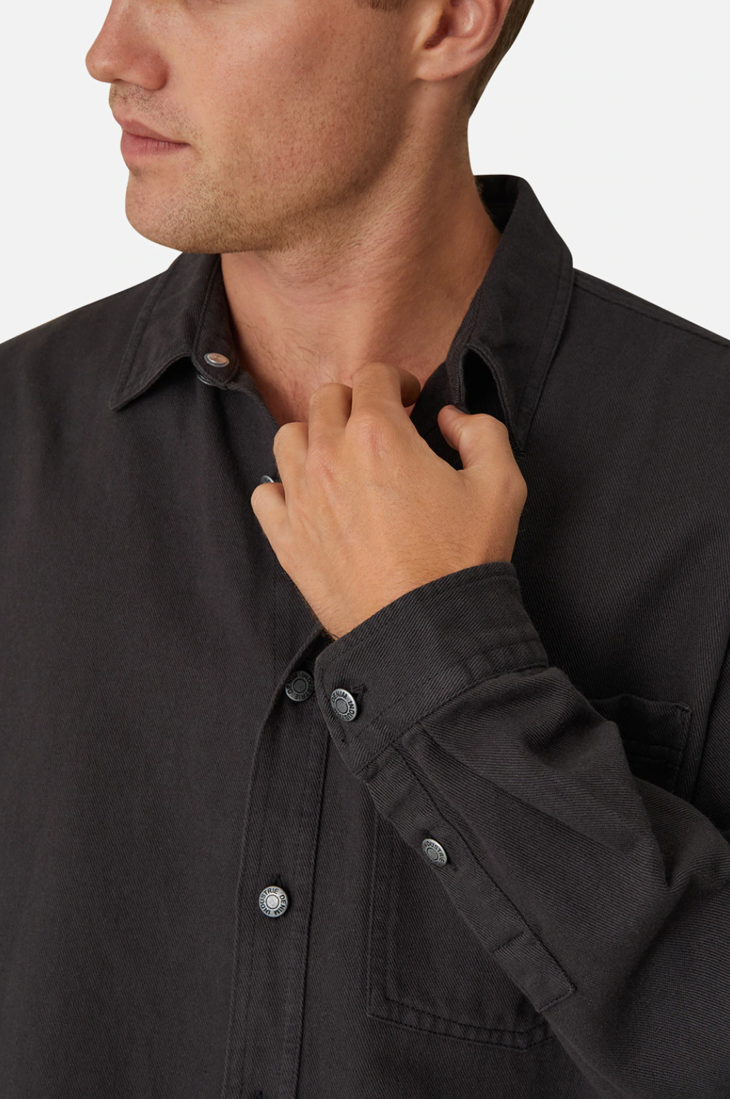 A person with short hair, partially visible, adjusts the collar of their stylish Industrie The Portland Shirt. The charcoal-colored cotton/linen blend shirt features a front pocket, and the person's face is turned slightly to the side, looking out of view.