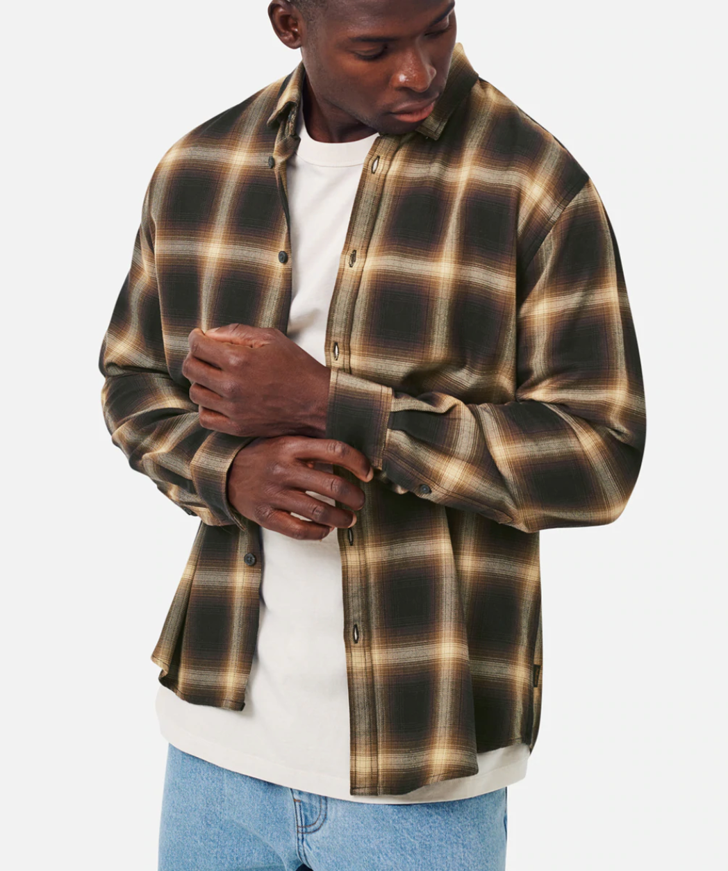 A person wearing the Industrie The Benson Shirt, a stylish and comfortable plaid design in brown, beige, and black hues over a white t-shirt, adjusts their sleeve while looking down. They stand against a plain white background paired with blue jeans.