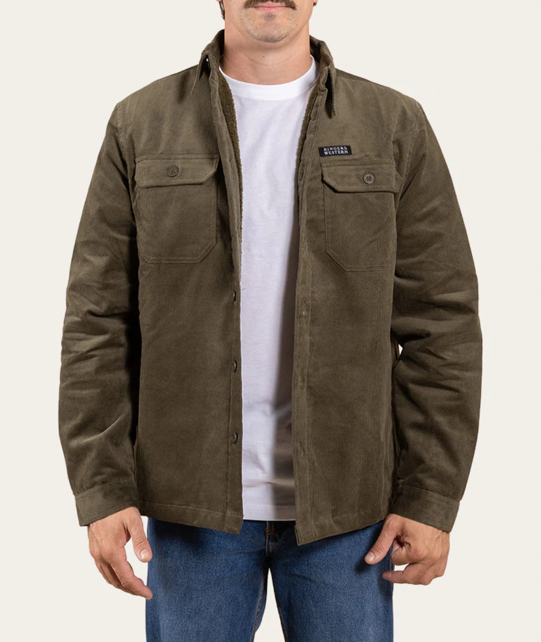 A man models the Ringers Western Ashwell Overshirt, a khaki, sherpa-lined jacket with two chest pockets, set against a plain background. Beneath it, he wears a white T-shirt and pairs it perfectly with blue jeans.