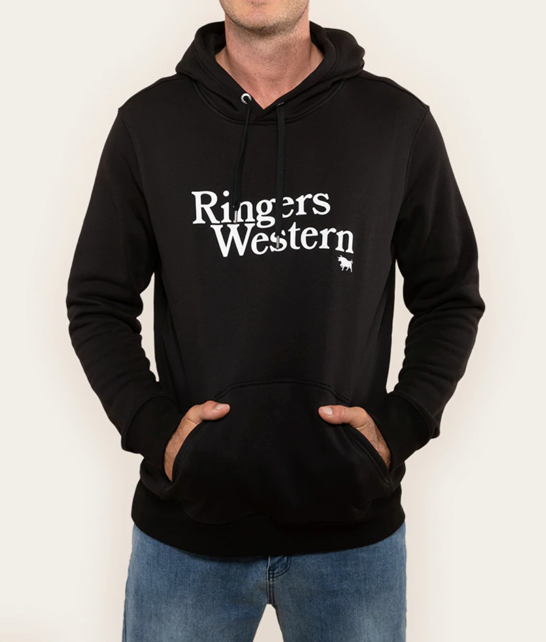 A person is dressed in a Ringers Western Lodge Hoodie, featuring a black color with "Ringers Western" printed in white on the front. This comfy hoodie includes a front pocket where the individual has both their hands tucked inside. Completing their look, they are wearing blue jeans against a plain, light-colored background.