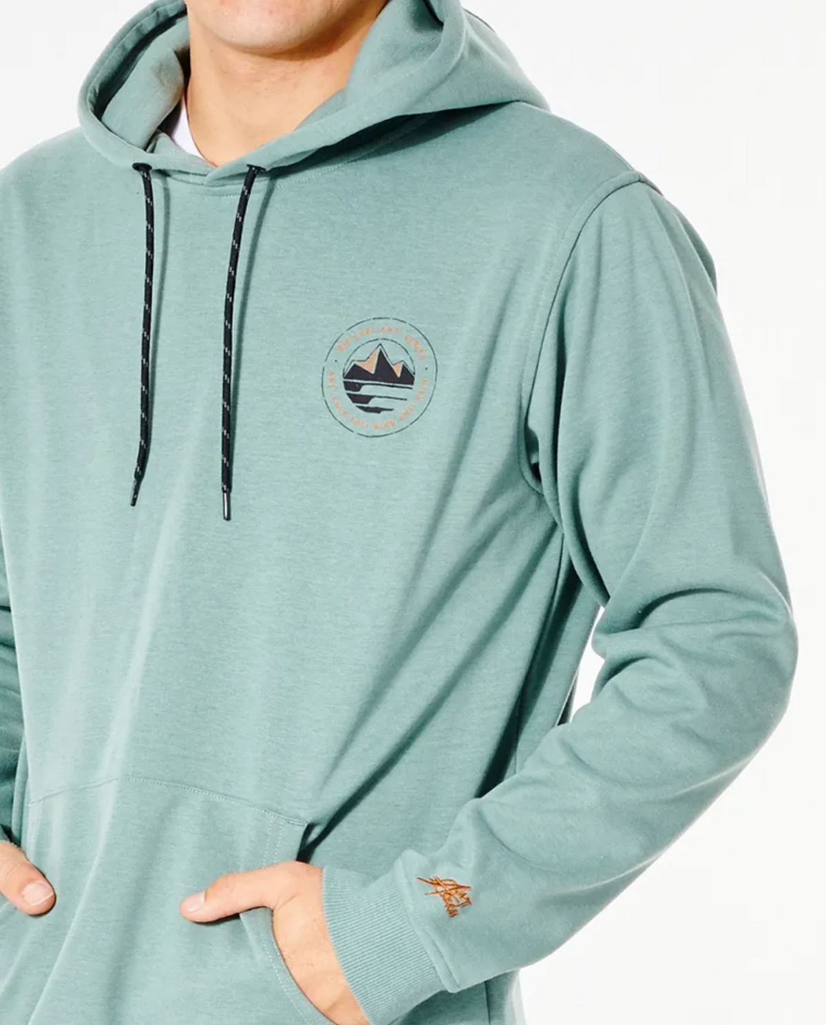 A person wearing the Rip Curl Anti Series Journey Hood in light teal, featuring a circular mountain and wave emblem. This winter hoodie has Durable Water Resistant properties, black drawstrings, and the individual has their hands in the front pocket. Only the upper body is visible.
