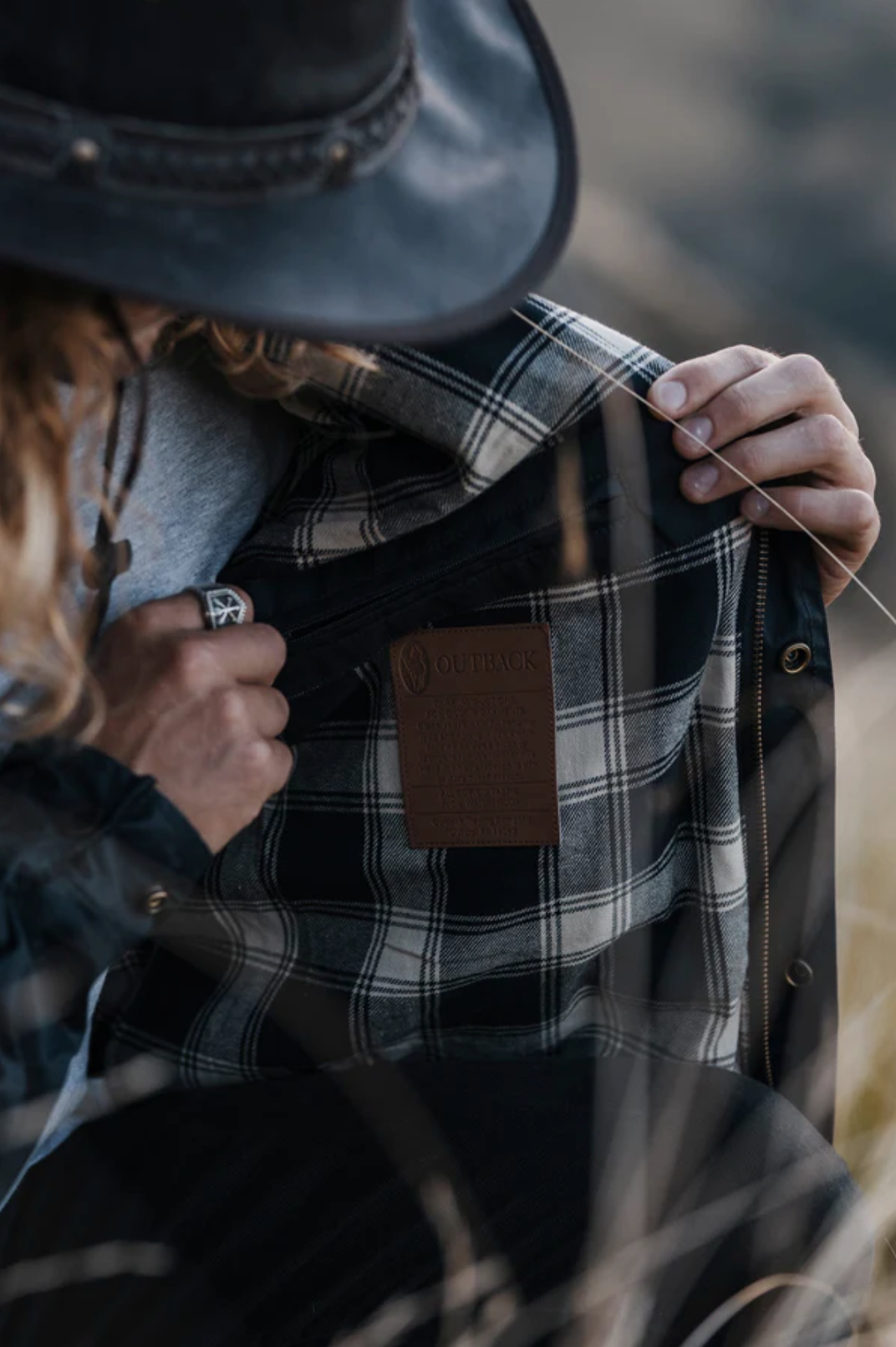 A person with long hair, wearing a dark hat and ring, holds open an Outback Burke Jacket showing its plaid flannel lining and brown label. The background features blurred natural tones that enhance the charm of this Australian waxed cotton waterproof jacket.