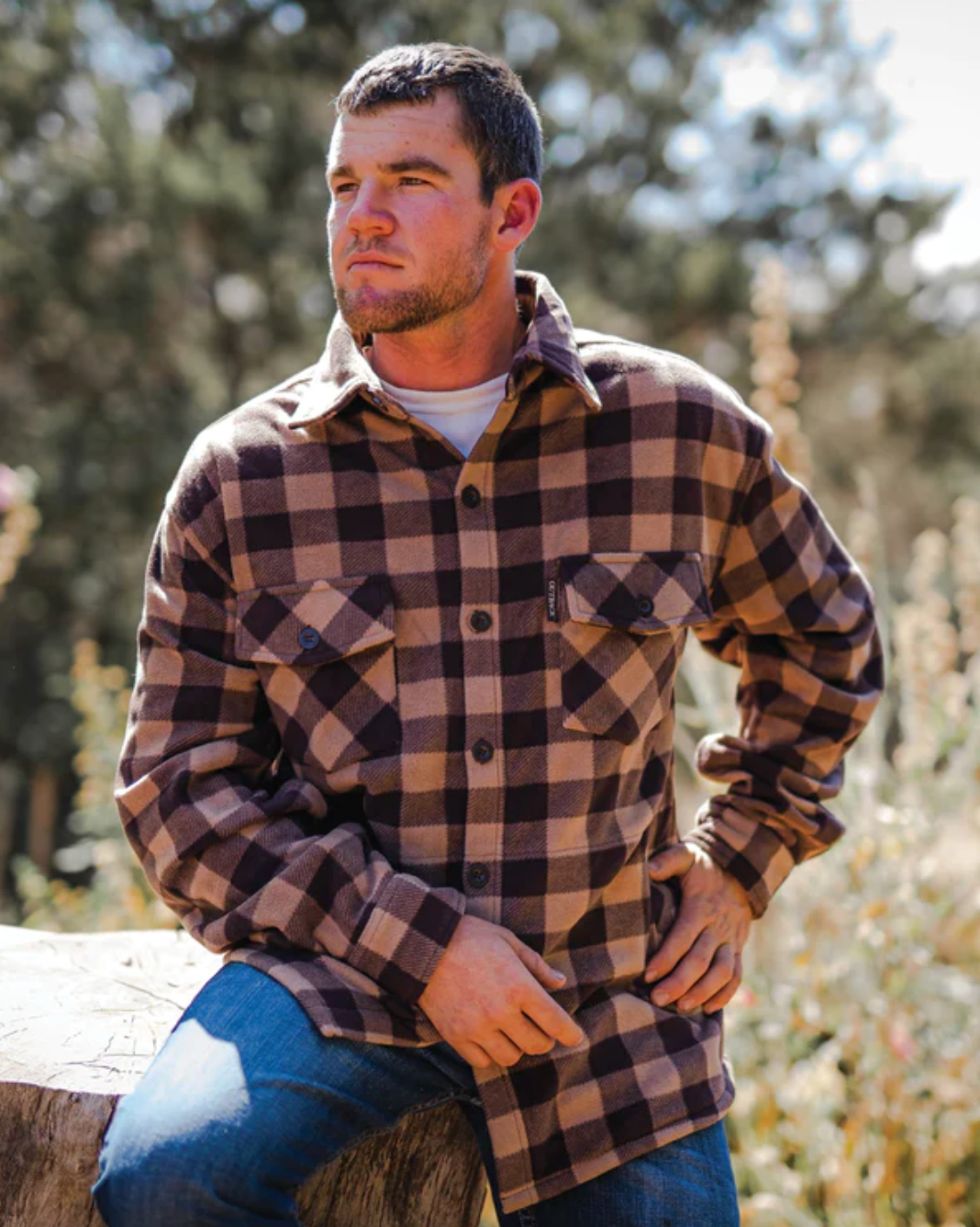 A man outdoors stands with one hand on his hip in an Outback Mens Big Shirt, a mid-weight garment featuring a cozy fleece design perfect for cool weather. Leaning against a large log, he looks to the right as sunlight filters through the leafy backdrop.