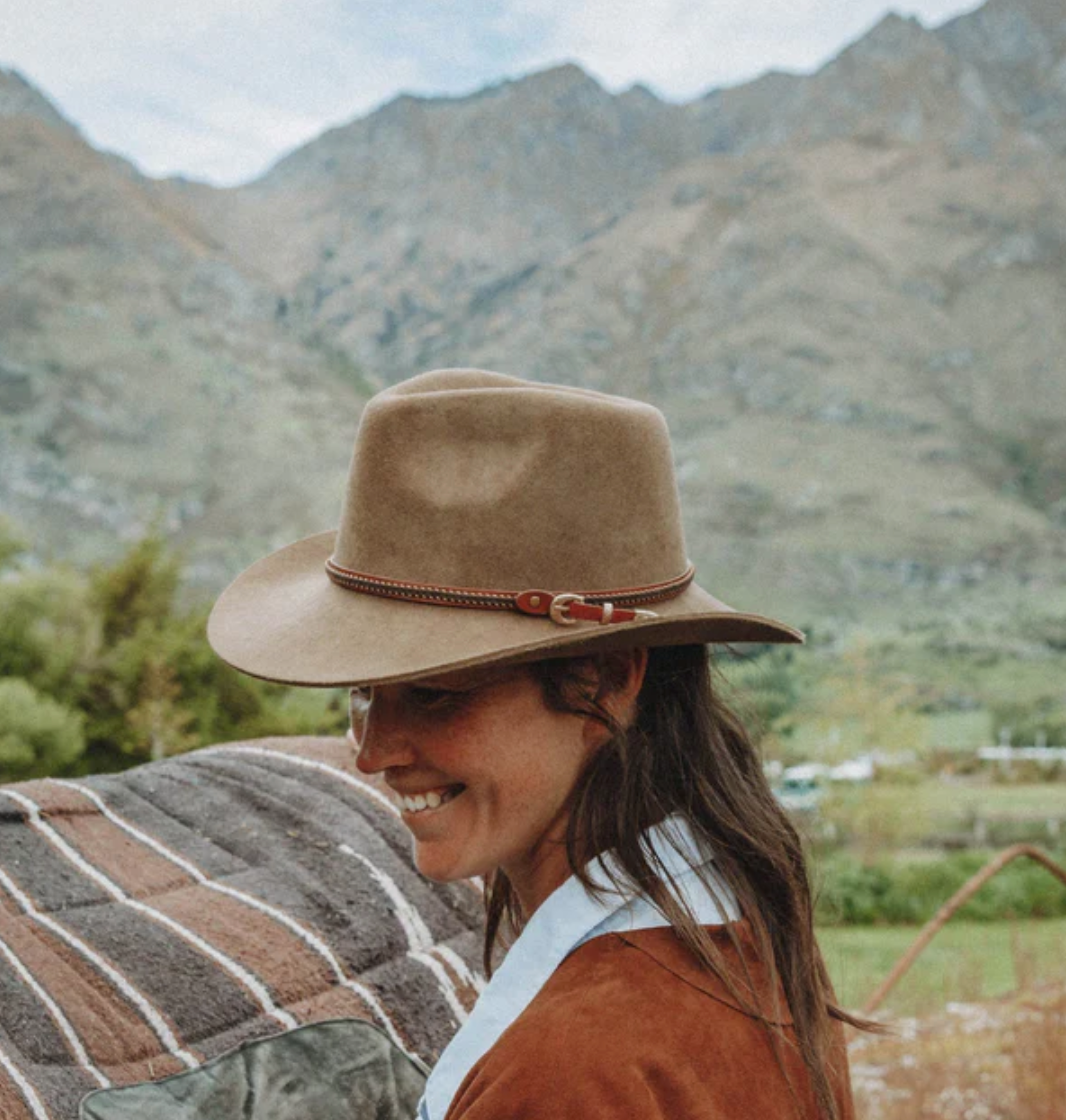 A woman wearing the Outback Nelson Wool Hat and a brown jacket smiles while standing next to a horse covered in a striped blanket. Mountains and greenery are visible in the background under a cloudy sky.