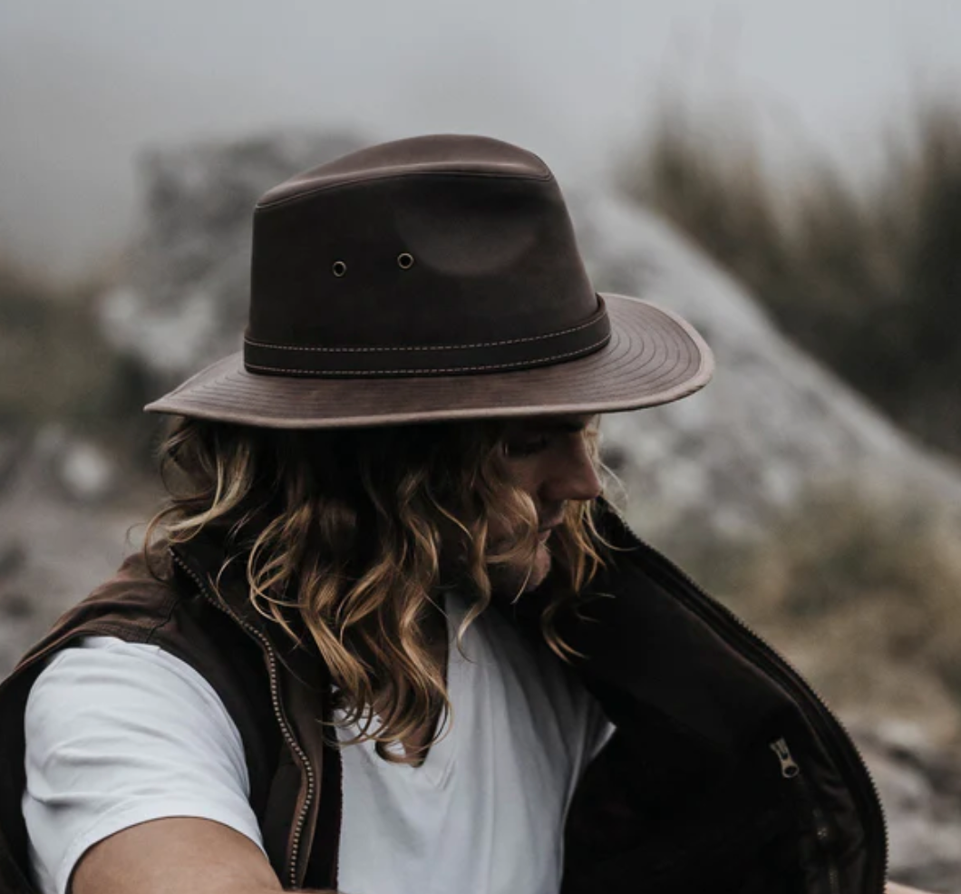 A person with long hair, dressed in a white shirt and brown vest, sits outdoors wearing the Outback Raven Hat. The wide-brimmed hat offers UPF protection and moisture-wicking properties, ensuring comfort as they look downward against a foggy backdrop of blurred rocks and grass.
