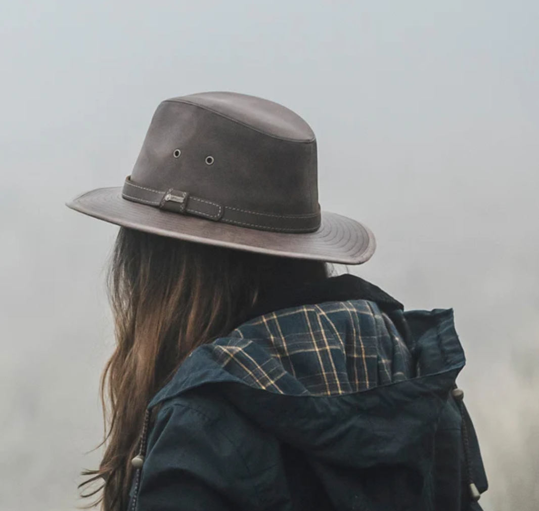 A person with long hair, wearing a blue plaid shirt and dark jacket, stands facing away from the camera, donning an Outback Raven Hat with a wide brim. The background appears foggy and indistinct.
