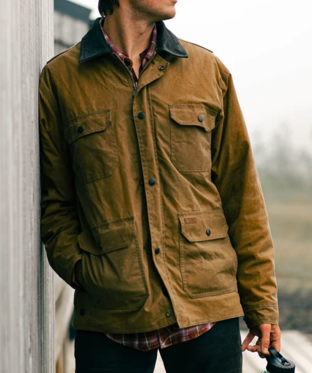 A person leans against a wooden wall, clad in the comfortable Outback Gidley Jacket made of brown waxed canvas, layered over a red plaid shirt. The waterproof jacket boasts multiple front pockets with buttons and dual entry pockets. The individual's face is not visible, and they are holding an object in their left hand.