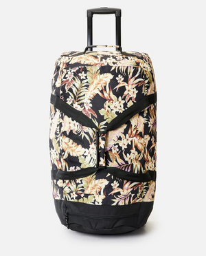The Rip Curl Jupiter 80L Sunday Swell Travel Bag is a spacious wheeled suitcase made from recycled polyester. It features a retractable handle and vibrant tropical floral design with light flowers on a black background, standing upright on a plain white surface.