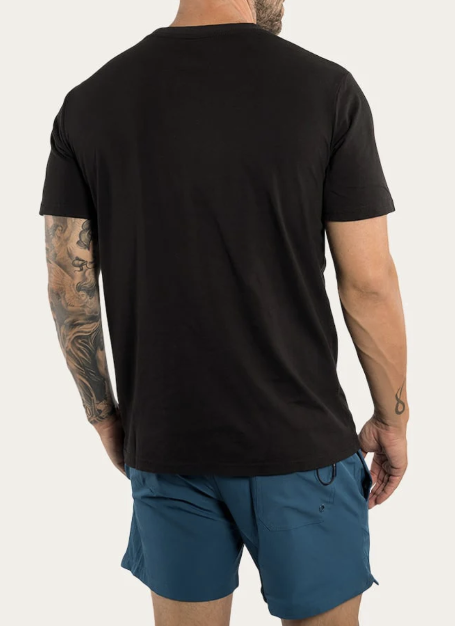 A person in a Ringers Western The Lodge Classic Fit T Shirt and blue shorts stands with their back to the camera. Tattoos adorn their right arm, and the 100% cotton tee complements the plain, light-colored background nicely.