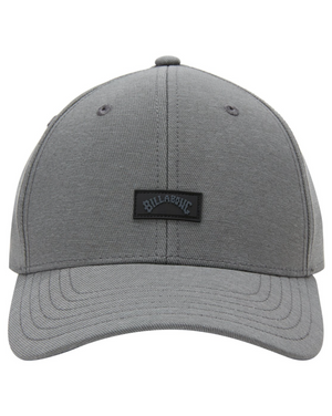 The Billabong Surftrek Snapback is a grey baseball cap with a rounded crown and slightly curved brim. It features a black rectangular patch on the front with "Billabong" in white lettering and is made from recycled polyester. Two small ventilation holes on the front panels add to its functional design.