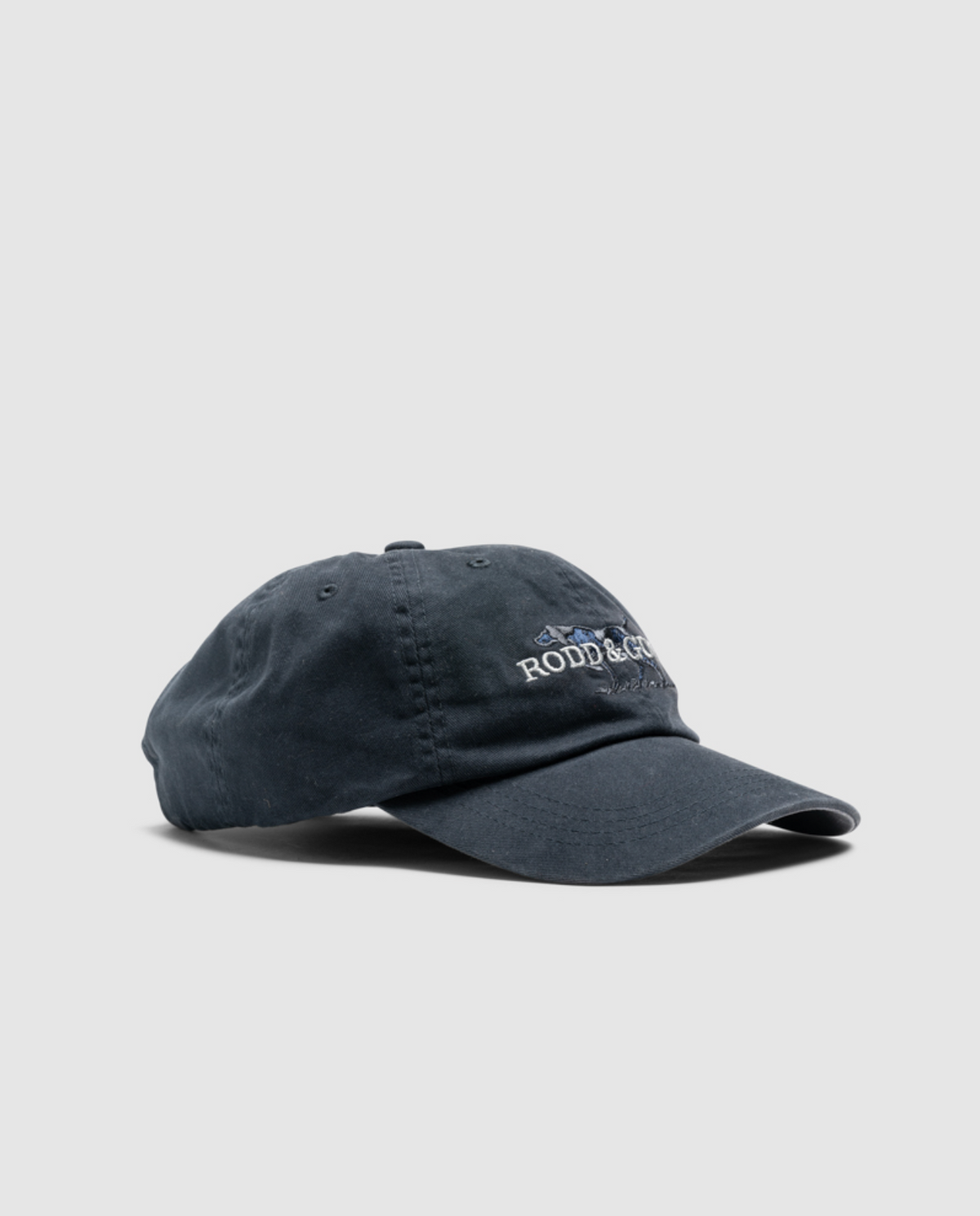 The Rodd & Gunn Double Barrel Cap in dark blue, made from sturdy cotton twill, has a slightly curved brim and features embroidered text and logo on the front, all set against a plain gray background for a casual worn look.