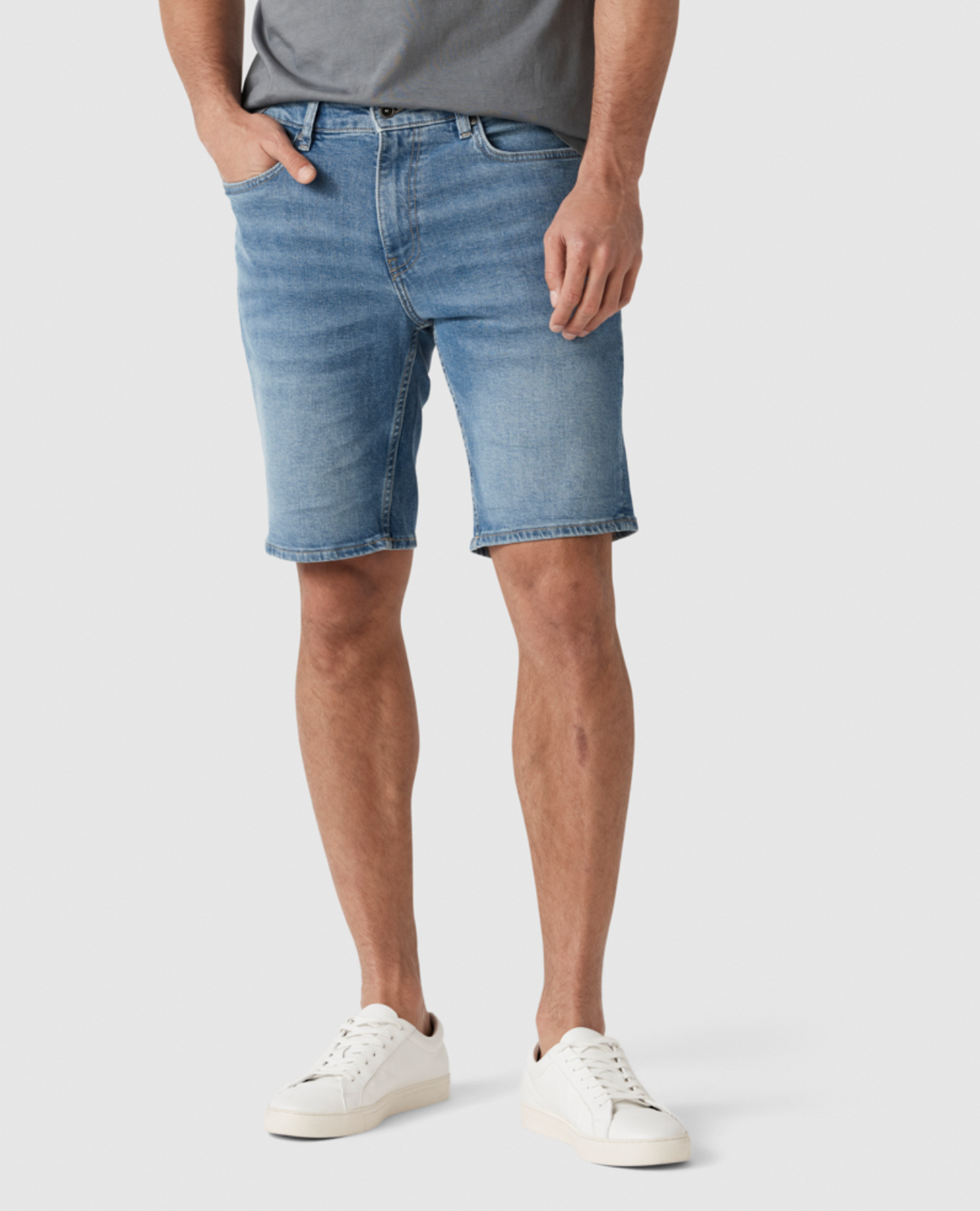 A person is standing against a plain background, wearing a grey t-shirt, the Rodd & Gunn Stephens Bay Summer Short in light blue with a mid-rise waist, and white sneakers. The person has their hands in their pockets, and only the lower half of their body is visible in the image.