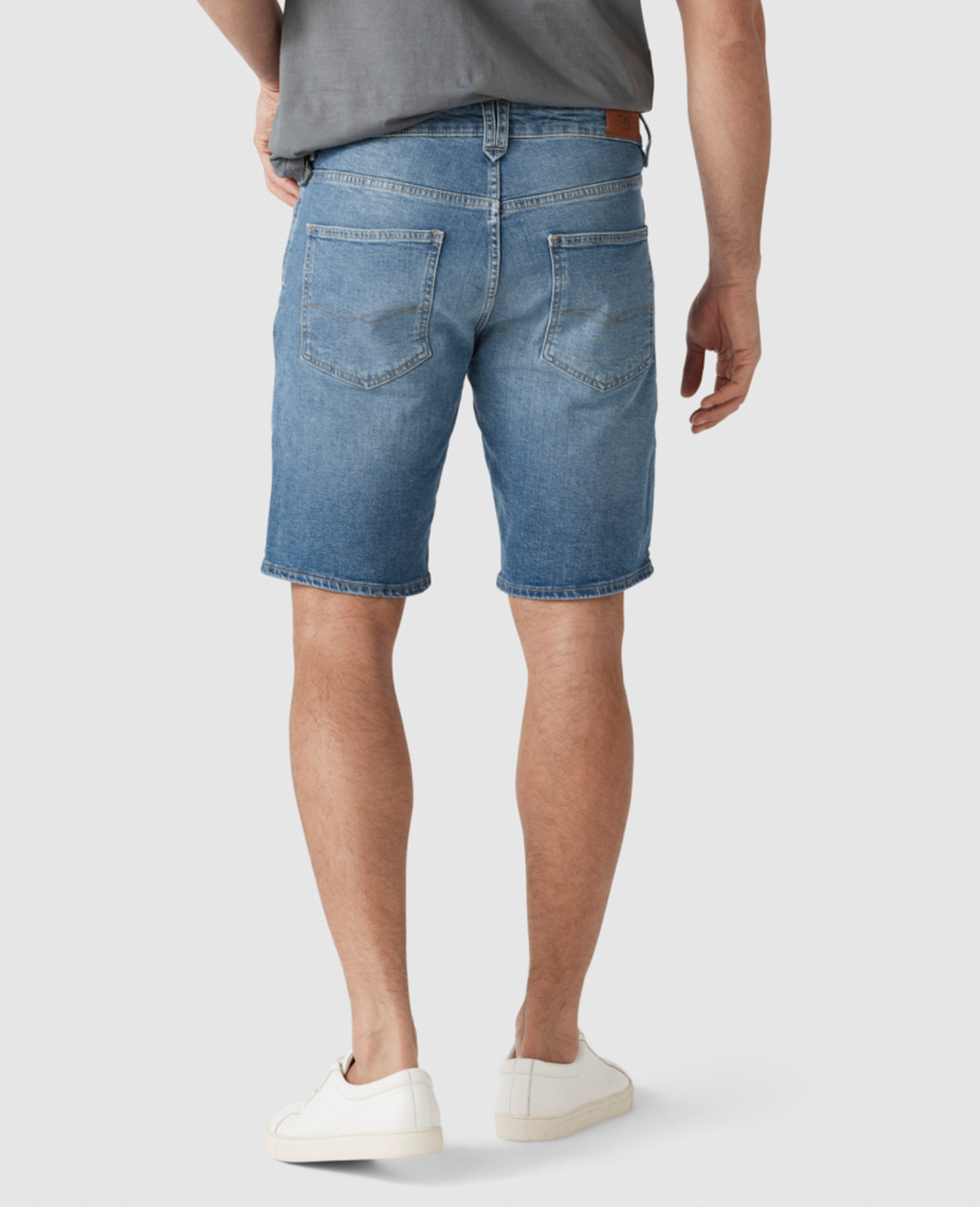 A person wearing the Rodd & Gunn Stephens Bay Summer Short in light blue, featuring a stretch fit and mid-rise waist, is standing with their back facing the camera. They are also wearing a gray T-shirt and white sneakers. The shorts have two back pockets and rolled-up hems, with the person's hands resting by their sides.