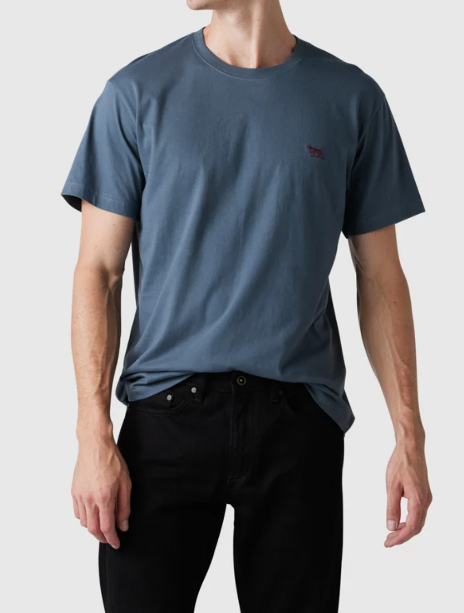 Someone stands against a grey background wearing The Gunn T-Shirt by Rodd & Gunn, a plain dark blue sports fit tee made from premium Australian cotton, featuring a small red logo on the chest. Their face is not visible.