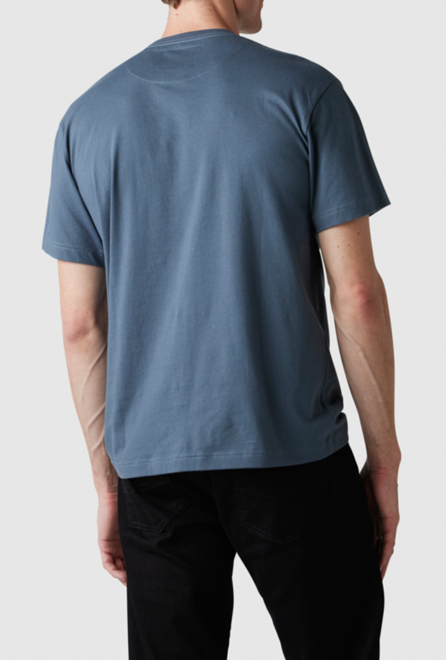 A person is standing with their back to the camera, wearing a Rodd & Gunn The Gunn T-Shirt in plain blue, made from soft Australian cotton, along with black pants. The background is a plain, light grey color.