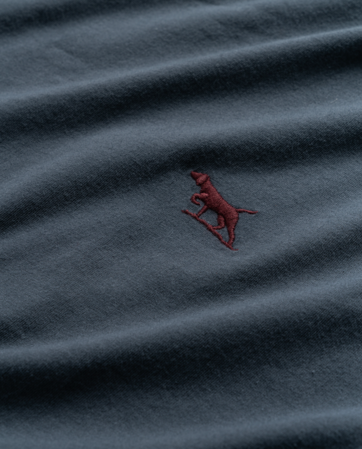 A close-up image of the Rodd & Gunn The Gunn T-Shirt, crafted from Australian cotton in a deep blue hue, features an embroidered logo of a red dog standing on its hind legs, seemingly holding or leaning on a small object. The fabric appears slightly creased.