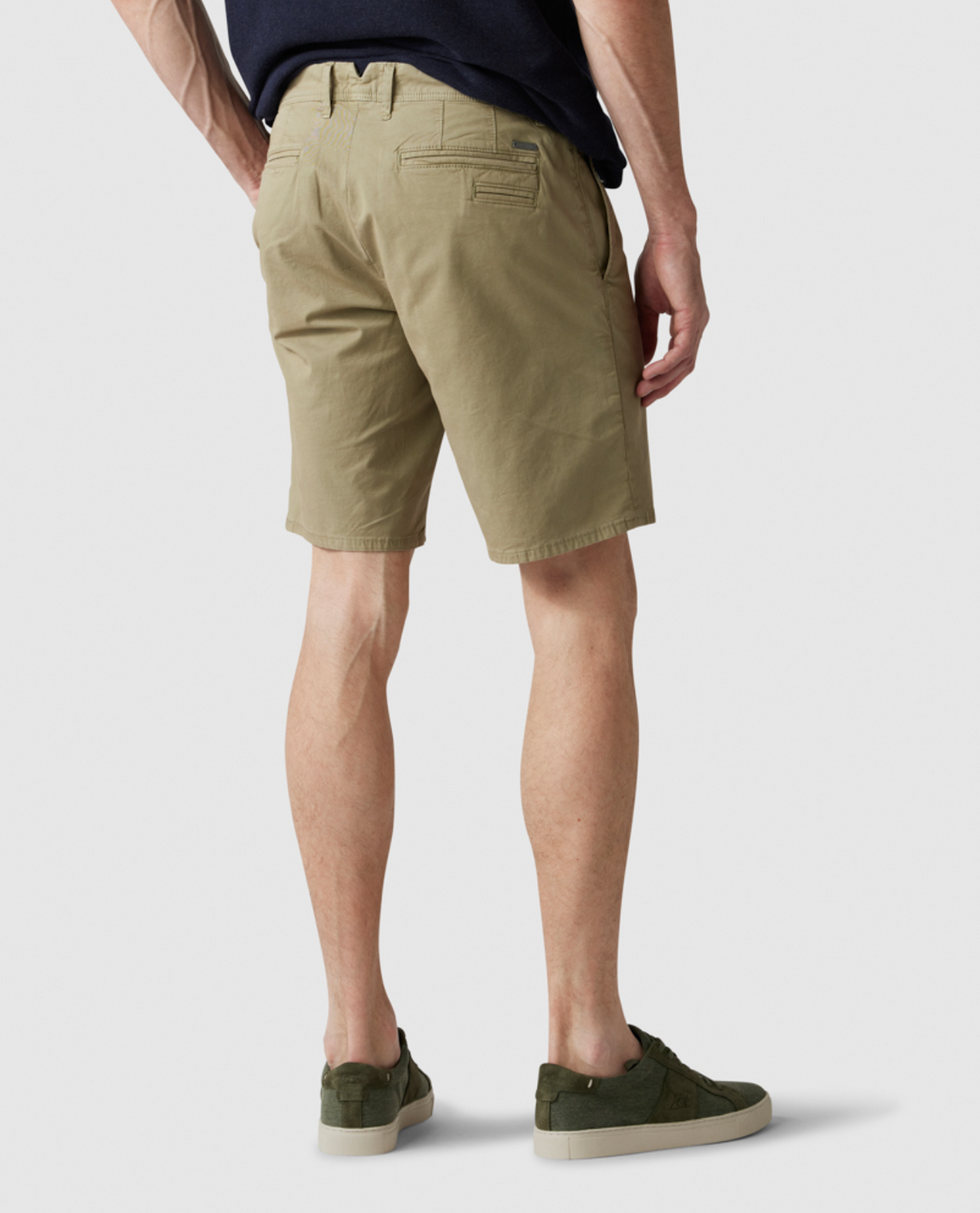 A person is shown from the mid-back to their legs, wearing Rodd & Gunn The Peaks Custom Shorts with pockets and a black belt, paired with a black shirt and green sneakers. The background is plain white, emphasizing the clothing. The person's stance is casual, with arms relaxed.