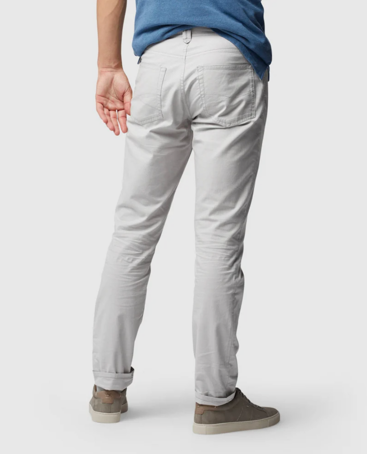 A rear view features someone in Rodd & Gunn Straight Fit Jeans with a mid-rise waist, light gray in color. They pair the jeans with a blue shirt and brown shoes with white soles, standing against a plain white background, placing one hand behind their back.