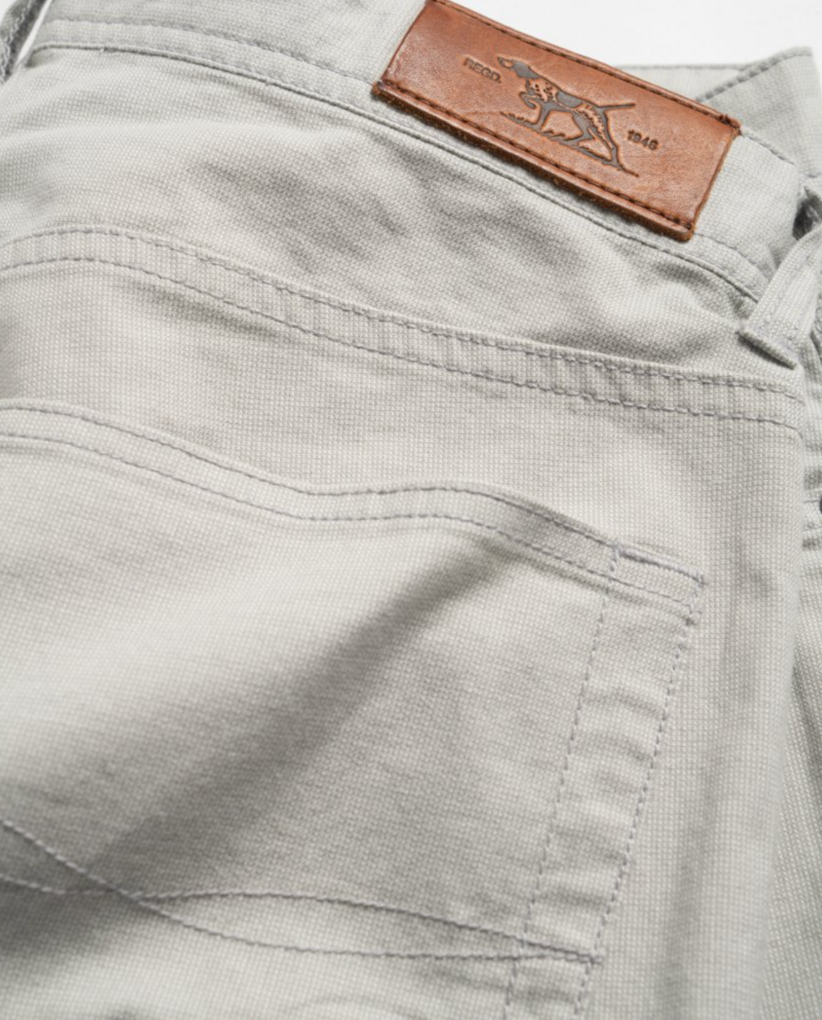 Close-up of Rodd & Gunn Straight Fit Jean in light gray, highlighting a back pocket with a brown leather patch displaying a stitched logo and silver rivets. The textured, durable fabric offers comfort with stretch fabric and features visible stitching around the pocket, belt loops, and mid-rise waist.