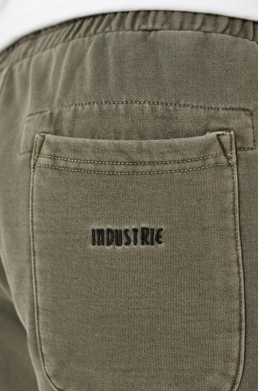 A close-up of the olive green Industrie The Del Sur Track Short reveals a pocket with "INDUSTRIE" embroidered above it in black thread. Its elastic waistband hints at an oversized fit, ideal for a relaxed track short style.