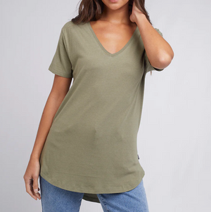 A person in a comfortable olive green Silent Theory Zuri Tee, made from soft cotton/bamboo jersey, and blue jeans stands against a plain white background, with their left hand on their head.