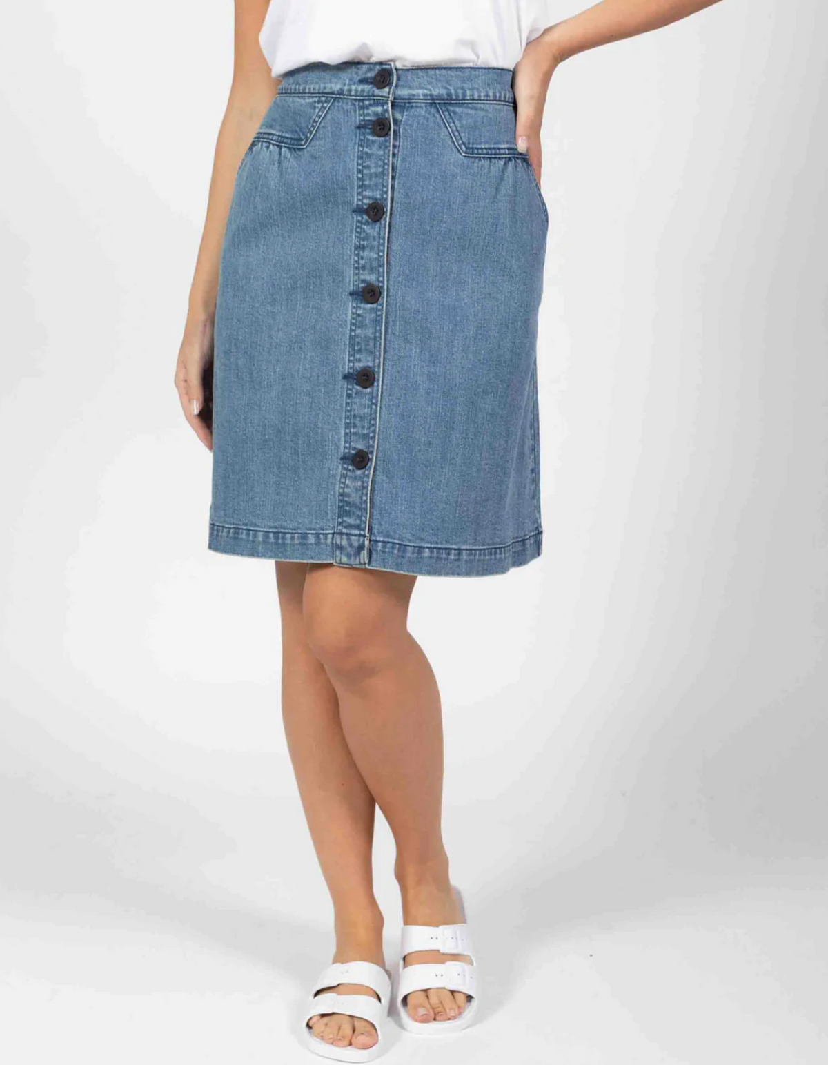 Wearing a white top and the Foxwood Amanda knee-length denim skirt featuring a button front and pockets, the person pairs this with white sandals. Their pose shows one hand resting on their hip against a plain background.