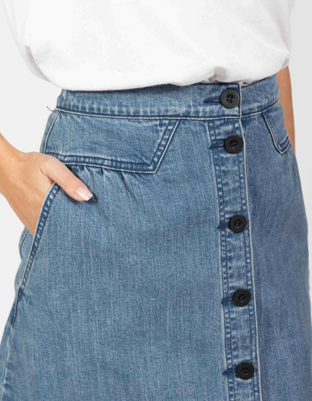 A close-up shows someone wearing the Foxwood Amanda Skirt, a high-waisted denim piece with an elastic waist and black button front. They have their hand in one of the side pockets, perfectly paired with a crisp white top.