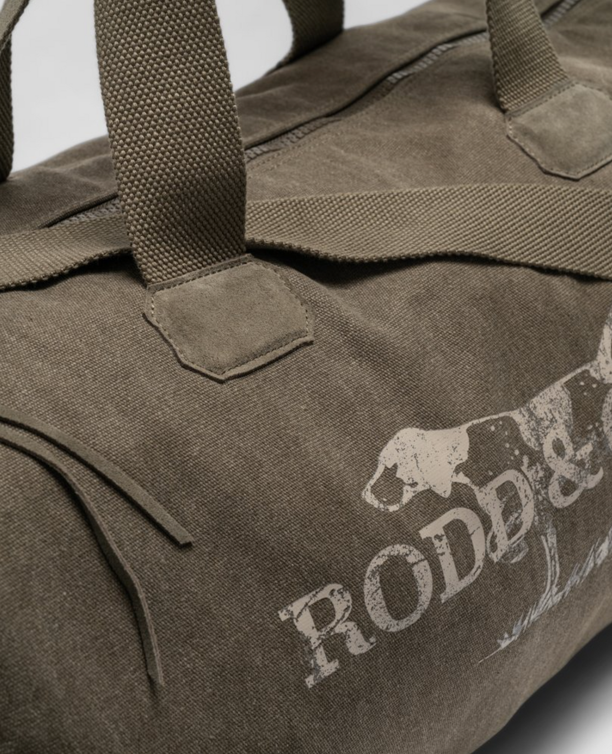 Close-up of a rugged brown cotton canvas Rodd & Gunn Double Barrel Duffle Bag with sturdy handles, featuring a partially visible dog print and "RODD & GUNN" text on the side.