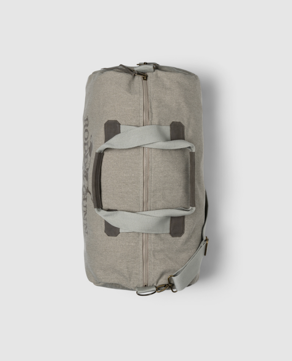 The Rodd & Gunn Double Barrel Duffle Bag is made from durable cotton canvas, featuring dark gray handles and a shoulder strap. This beige bag, with Rodd & Gunn branding, includes two brown accent straps across the middle on a light gray background.