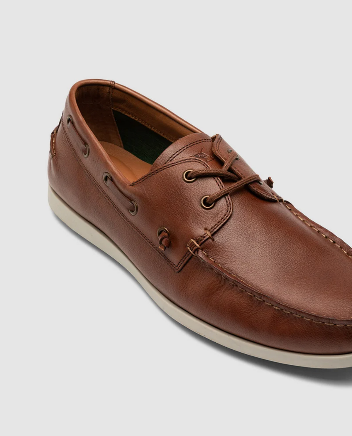 The Rodd & Gunn Gordons Bay Boat Shoe features a brown leather upper with laces, stitching details, metal eyelets, and a white rubber outsole, all set against a plain gray background.