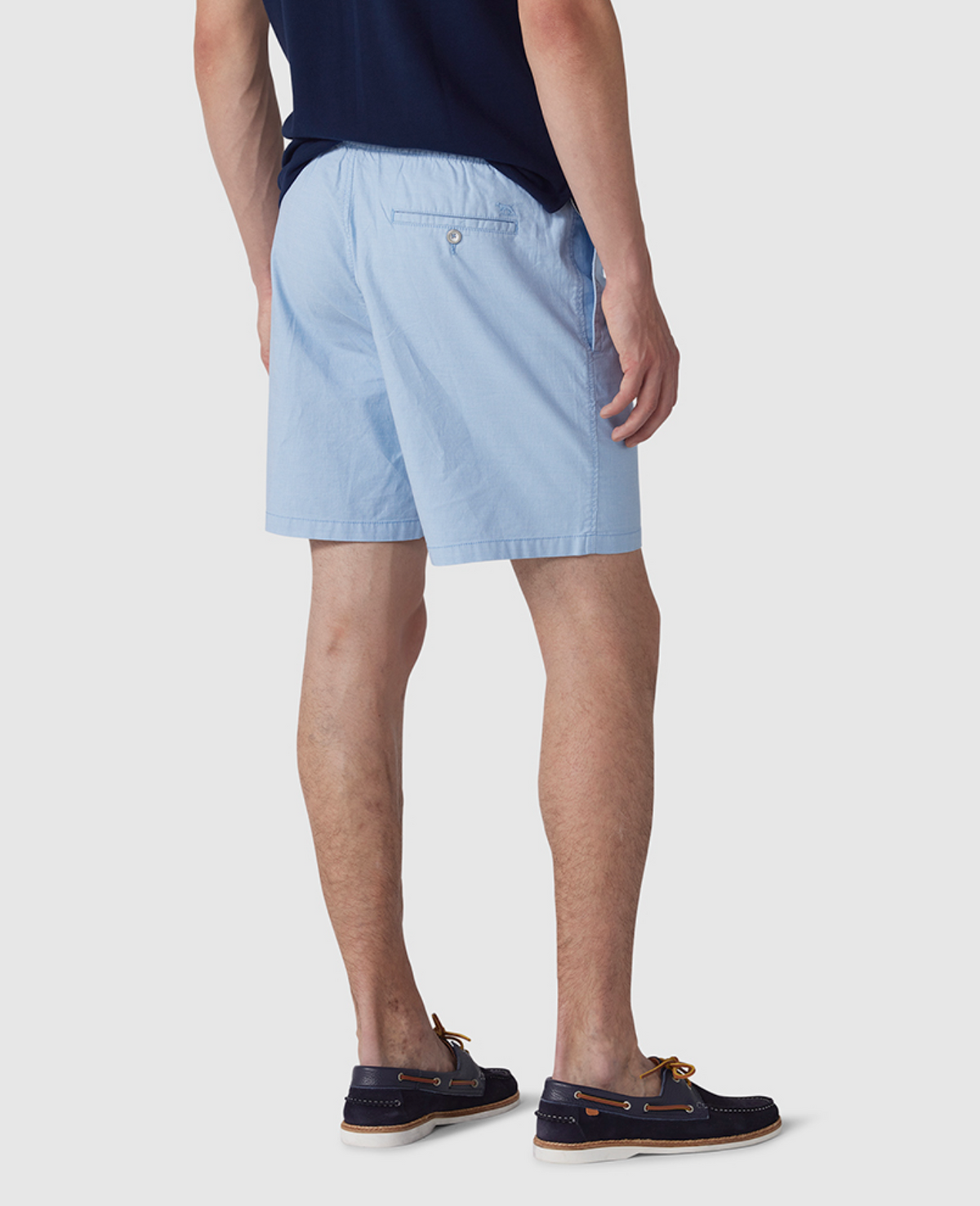 A person is seen from behind wearing Rodd & Gunn Resort Shorts with an elastic waist, a dark navy shirt, and navy boat shoes with tan laces against a plain gray background.