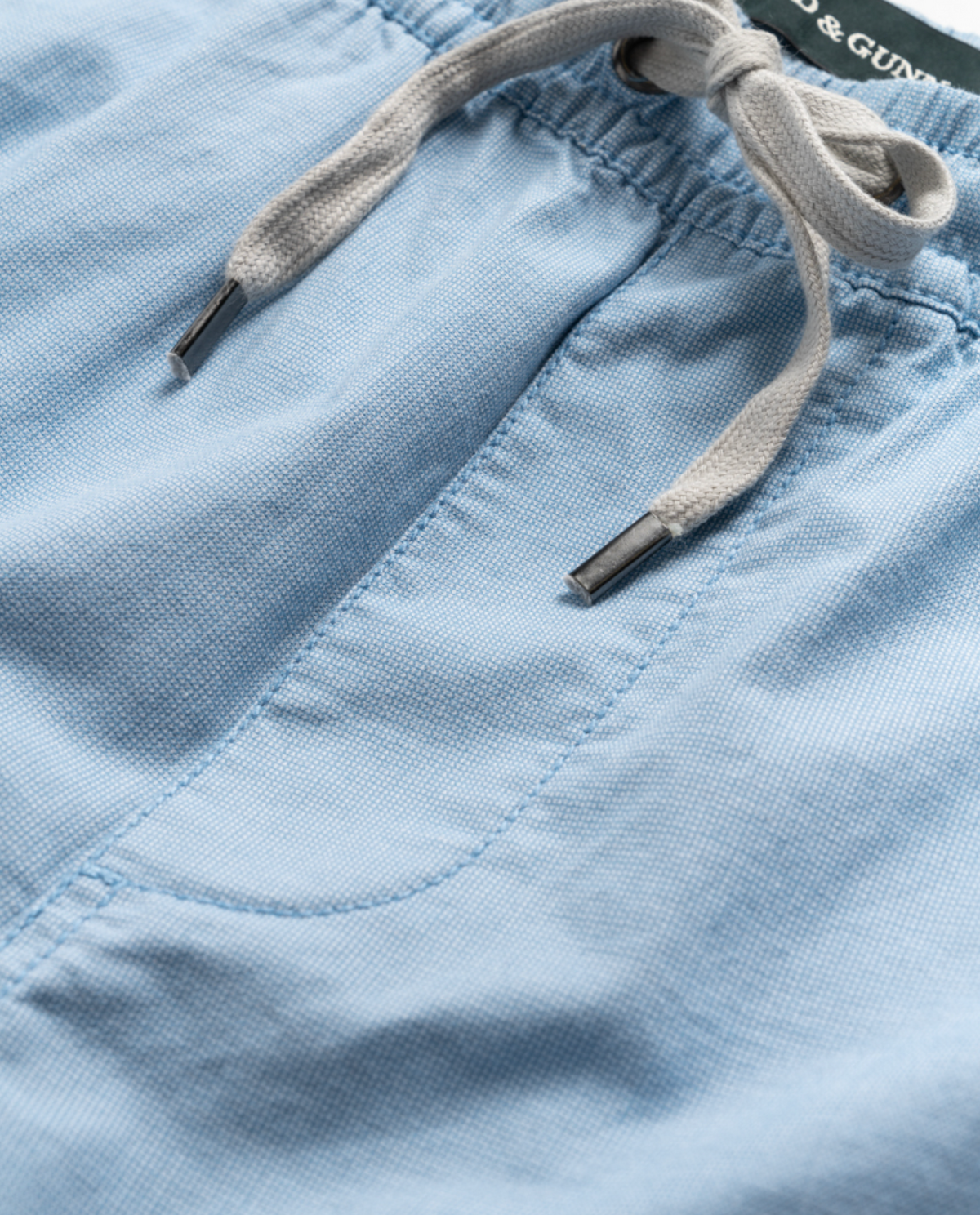 Close-up of light blue Rodd & Gunn Resort Shorts, made from cotton elastane with a fabric texture. They feature a gray drawstring with metal tips at the elastic waist for flexibility, and visible stitching detail that highlights their craftsmanship.