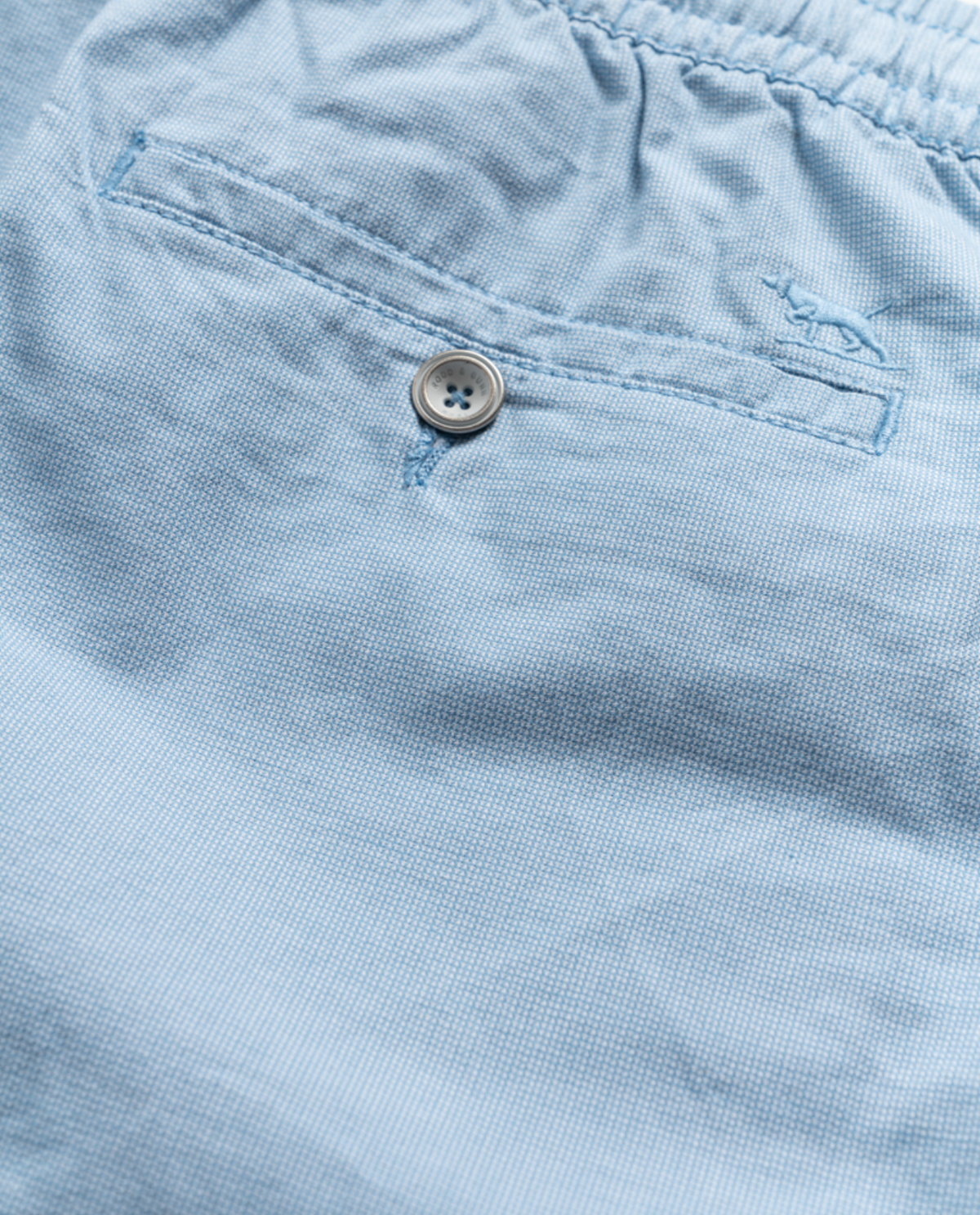 A close-up of the Rodd & Gunn Resort Short in light blue cotton elastane showcases a buttoned pocket with an embroidered logo. The fabric texture is slightly textured with visible stitching detail, exemplifying Rodd & Gunn shorts craftsmanship.
