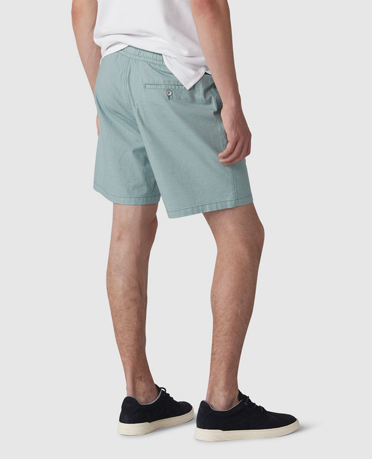 A person in a white T-shirt, Rodd & Gunn Resort Shorts in light teal with an elastic waist, and black sneakers with white soles stands against a plain white background, partially turned away from the camera.